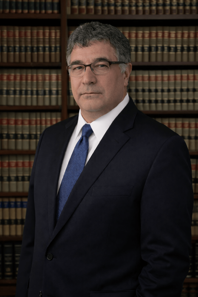 A middle-aged man in a dark suit stands confidently in front of a bookcase filled with law books, symbolizing legal expertise and professionalism.